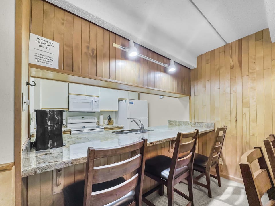 A small kitchen with wooden cabinets and a marble countertop, equipped with an oven, microwave, refrigerator, and three wooden bar stools in front. A sign with emergency contact information is on the wall.