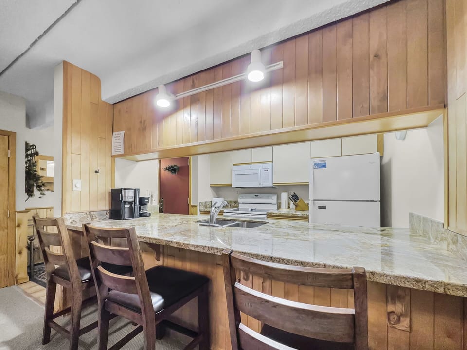 A kitchen with wooden cabinets and a marble countertop, containing three barstools, a coffee maker, a microwave, a stove, and a refrigerator.