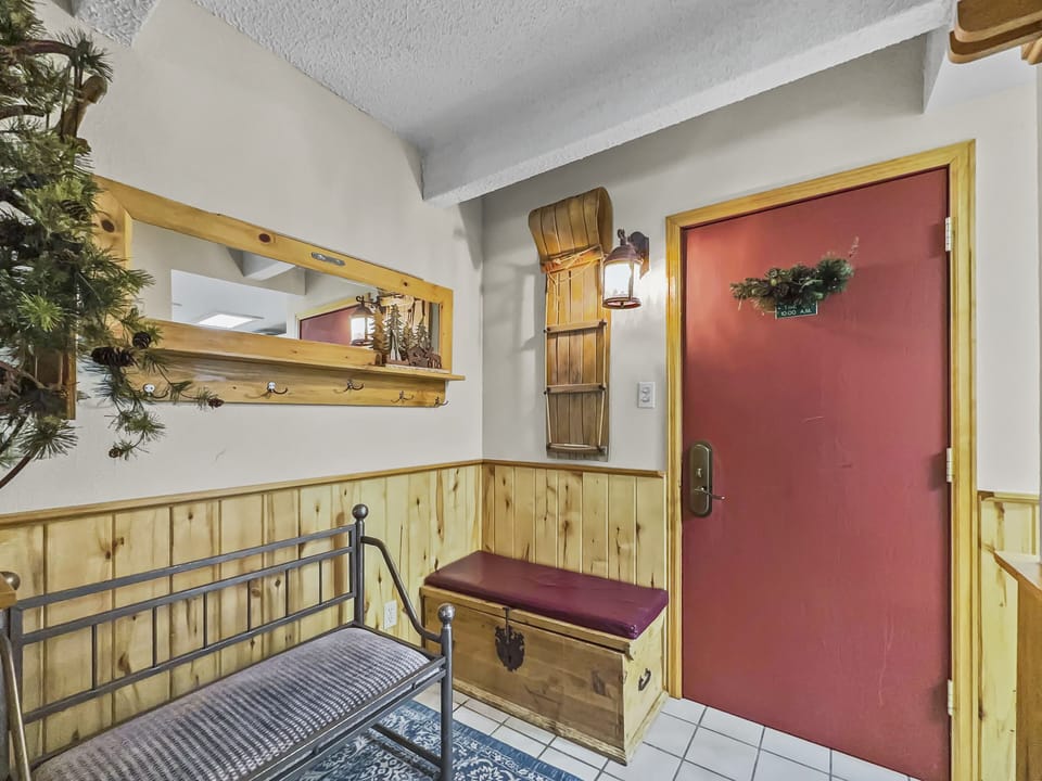 Small entryway with red door, wooden bench with red cushion, and decorative plants. A wall-mounted mirror with coat hooks is above the bench, next to a lantern-style light fixture.