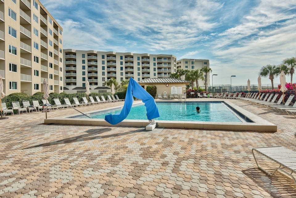 Outdoor swimming pool surrounded by multi-story resort buildings and palm trees under partly cloudy skies.