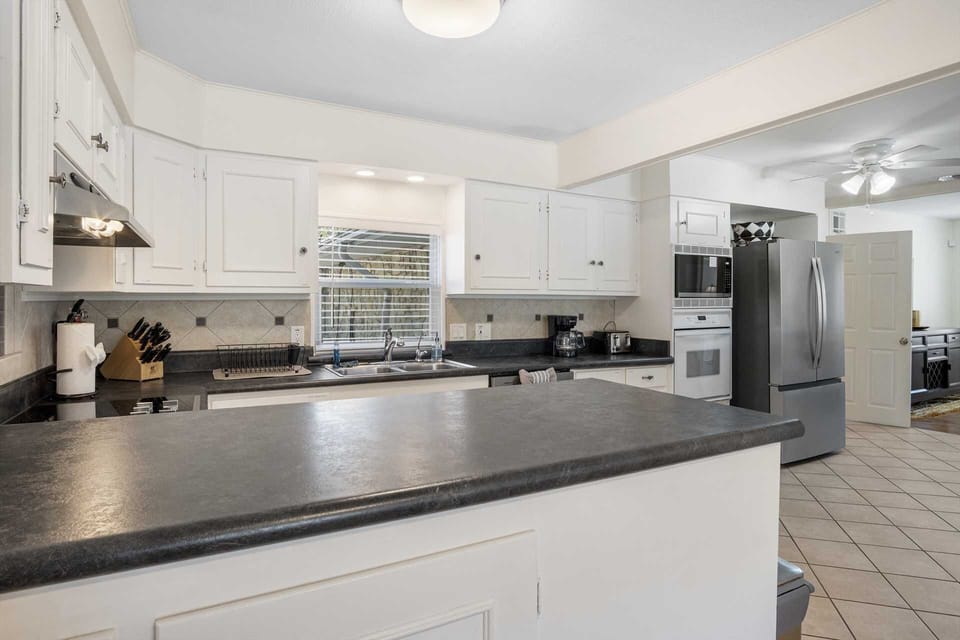 A clean, well-lit kitchen featuring white cabinetry, dark grey countertops, and stainless steel appliances, including a French-door refrigerator and built-in wall oven.