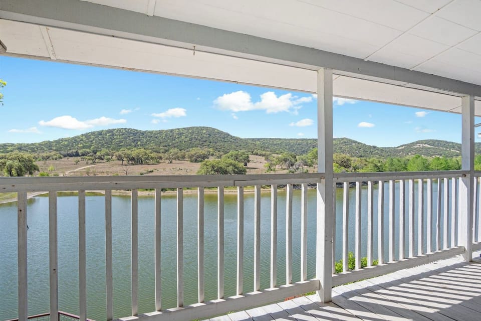 A stunning view from the private covered balcony, overlooking a peaceful body of water and the rolling, tree-covered hills of the Texas landscape under a clear blue sky.
