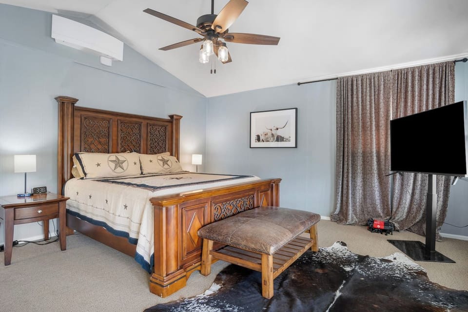 A spacious primary bedroom with a rustic Texas-themed aesthetic, featuring an ornate wood-carved bed frame, a leather-topped bench, and a large cowhide rug over neutral carpeting.