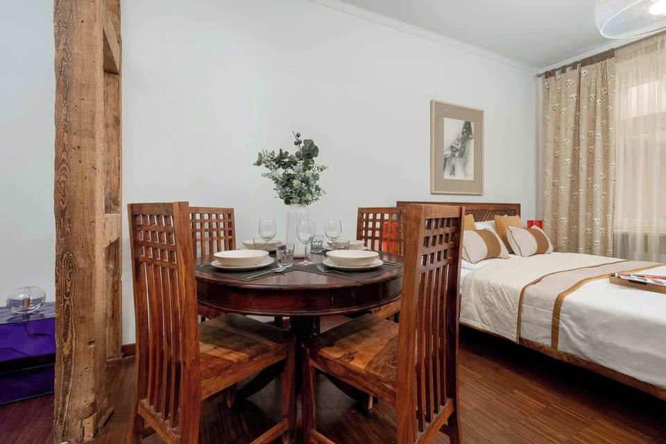 A round wooden dining table stands near a bed in a bright studio room. Soft curtains and light walls create a calm and peaceful setting.