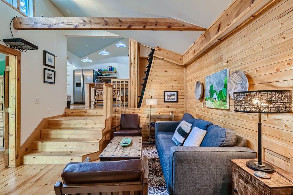 A cozy wooden interior with an open loft design featuring a timber staircase, a gray sofa, a rustic coffee table, a lamp, and wall art. The kitchen area is visible in the background.