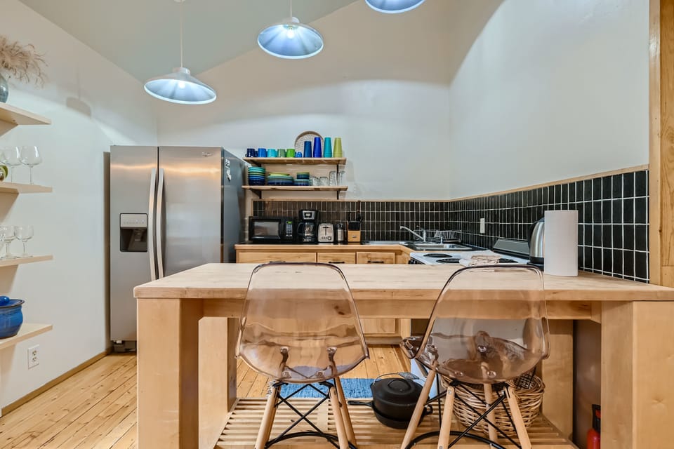 A modern kitchen with wooden countertops, black backsplash, stainless steel appliances, and clear plastic chairs at an island. Shelves with dishes and glasses are on the left wall.