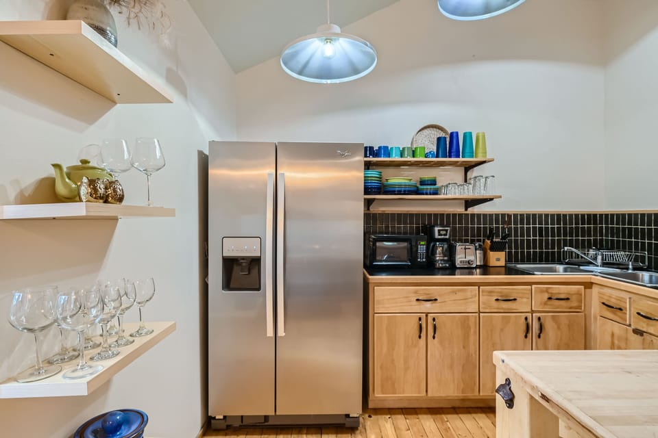 Modern kitchen with stainless steel refrigerator, wooden cabinets, and open shelving. Shelves hold glassware and tableware. Countertop features a microwave, coffee maker, and other kitchen essentials.