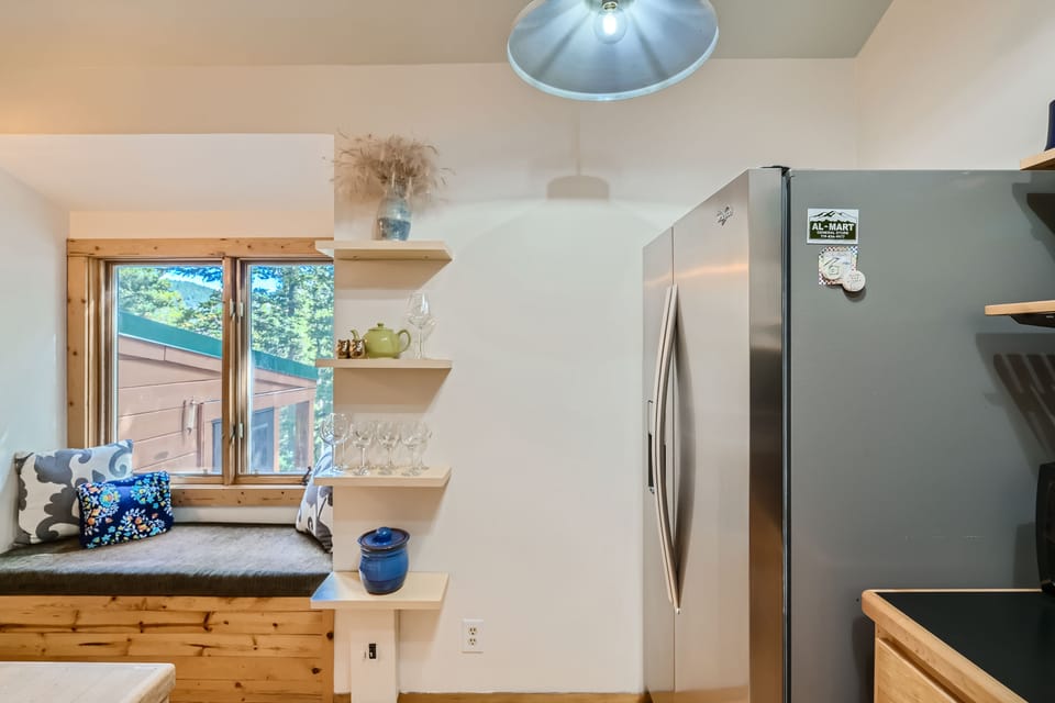 A kitchen corner featuring a stainless steel refrigerator, wooden shelves with glasses, a ceramic pitcher, and a blue vase. A window seat with cushions is visible beside the shelves.