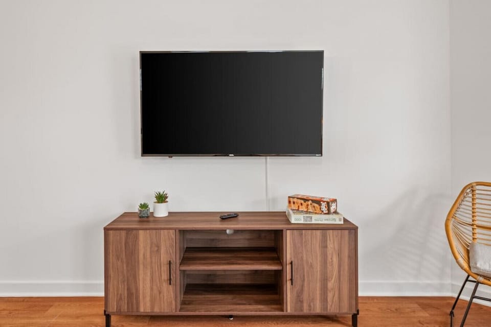 Bright and open living room filled with natural light, featuring a plush sofa, wall‑mounted Roku TV, ceiling fan, and workspace. The open‑concept layout flows seamlessly into the dining area and kitchen, blending historic charm with modern comfort.