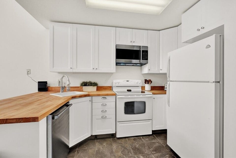 Clean and practical kitchen featuring full-size appliances, white cabinetry, microwave, oven, refrigerator, and warm wood countertops.