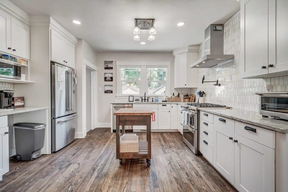 Clean, white kitchen with stainless appliances, open counters, and a central island with seating. Cook meals, prep snacks, or linger over coffee while the wide layout and wood floors keep everything feeling open and inviting.