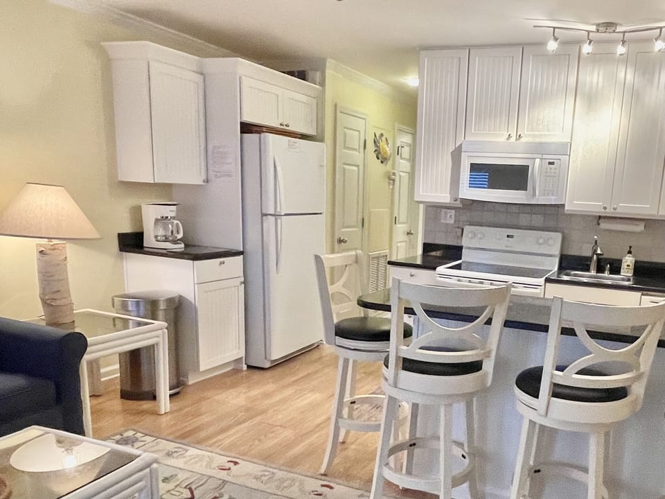 Compact kitchen with white cabinets, refrigerator, stove, and microwave. A small island with bar stools is in the foreground, adjacent to a living area with a lamp and side table.