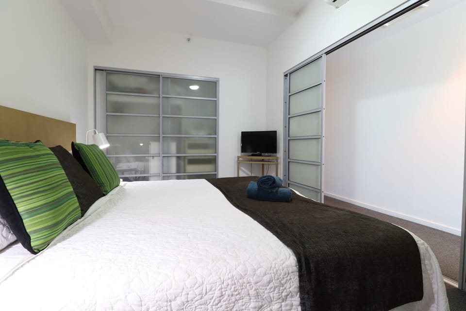 A restful bedroom with a large bed, crisp white quilted linens, and a soft dark throw. Frosted sliding wardrobe doors offer storage for longer stays, and a TV on a small desk makes it easy to unwind at the end of the day.