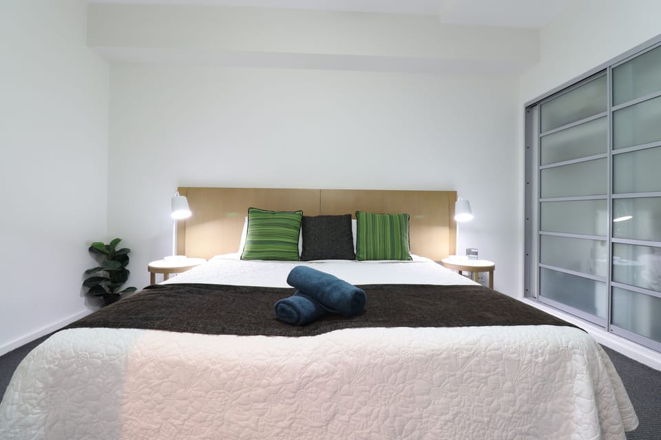 A calm bedroom with a large bed, crisp white linens, and green striped cushions against a warm timber headboard. Bedside tables with reading lamps on both sides make it easy to wind down and rest well after a full day.