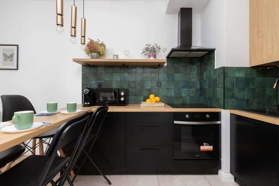 A modern kitchen with green tiled backsplash, black cabinets, and a dining table with black chairs.
