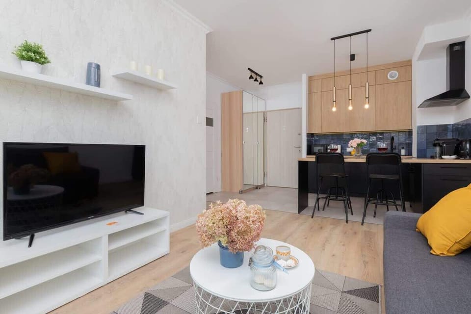 A wide-angle view of a living room with neutral tones, stylish furniture, and a kitchen visible in the background.