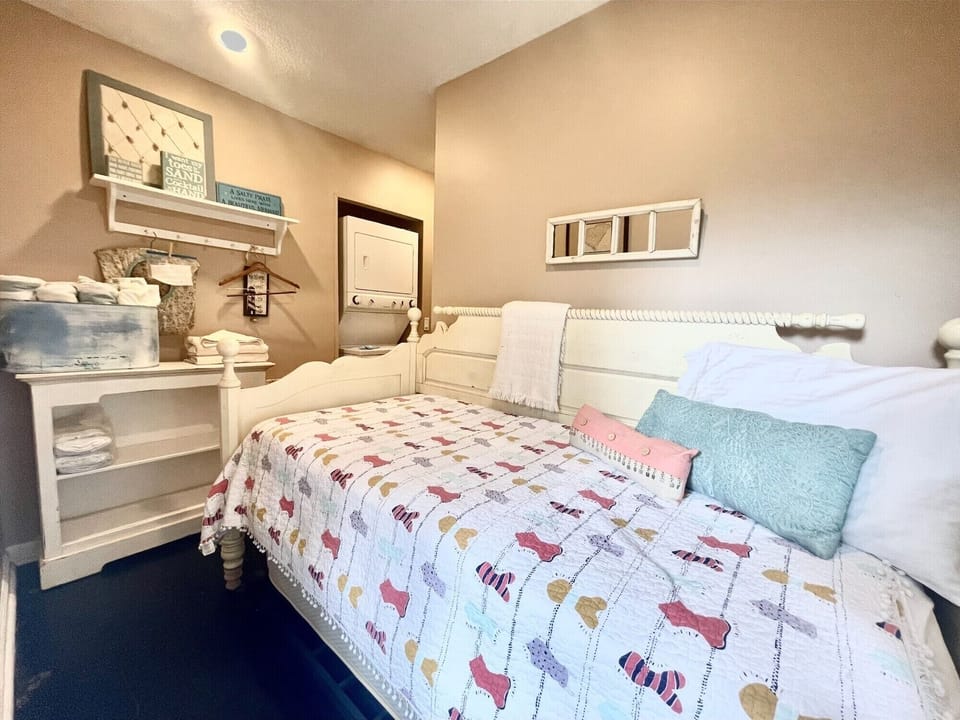 A cozy bedroom with a twin bed featuring a colorful quilt, multiple pillows, and a shelving unit with towels and decor. A stackable washer and dryer are visible in the corner.