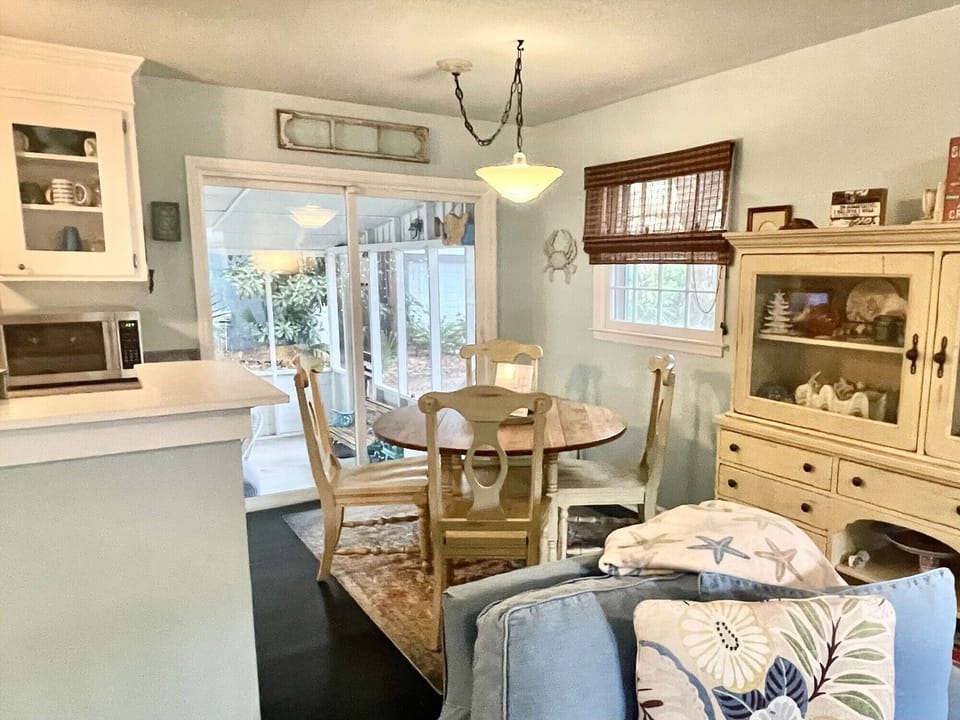 A cozy dining area with a round wooden table and four chairs near a window. A light blue couch with patterned pillows is in the foreground, and a hutch displays decorative items.