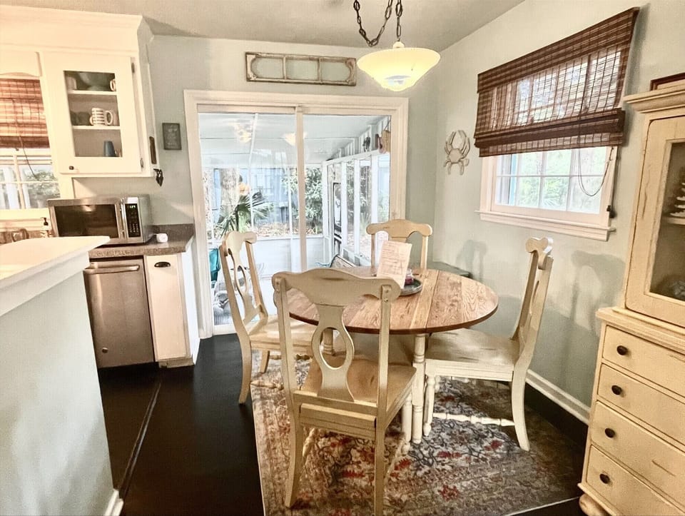 A cozy dining area with a round wooden table and four chairs, adjacent to a kitchen with stainless steel appliances. A large window opens to a sunlit porch. Cream and pastel decor create a warm atmosphere.