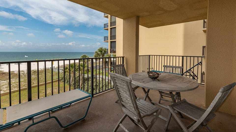 The balcony features a round wooden table with chairs, offering a serene view of the beach and calm waters beyond