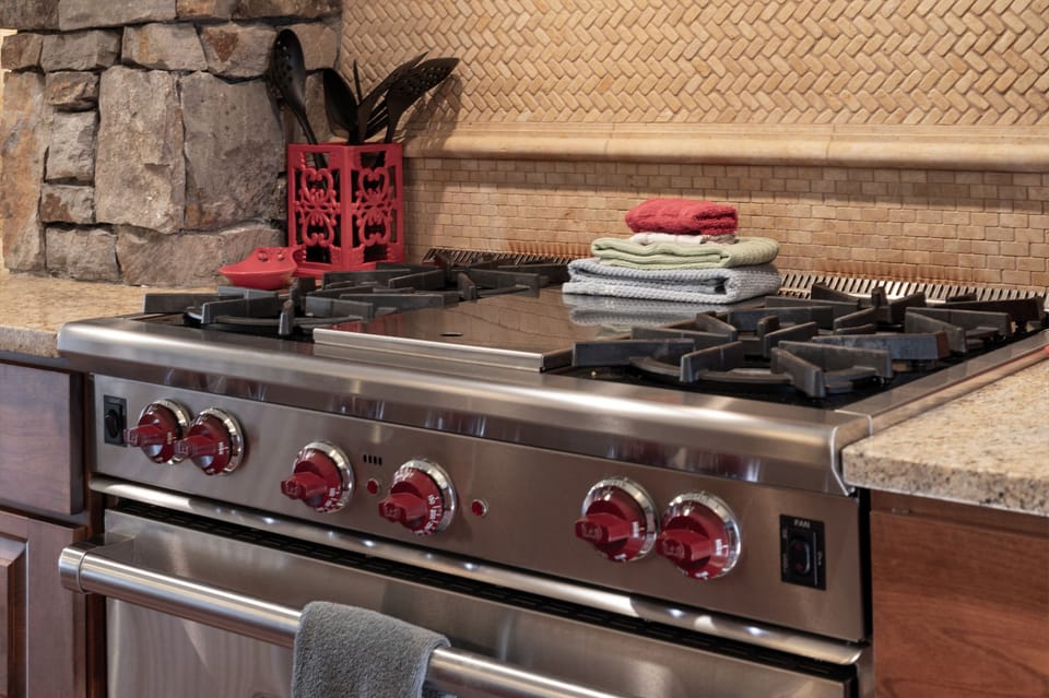 - Professional grade gas stove with red knobs and four burners for gourmet cooking - Handy utensils and folded dish towels keep everything within easy reach - Textured backsplash and stone accents complete the rustic kitchen aesthetic