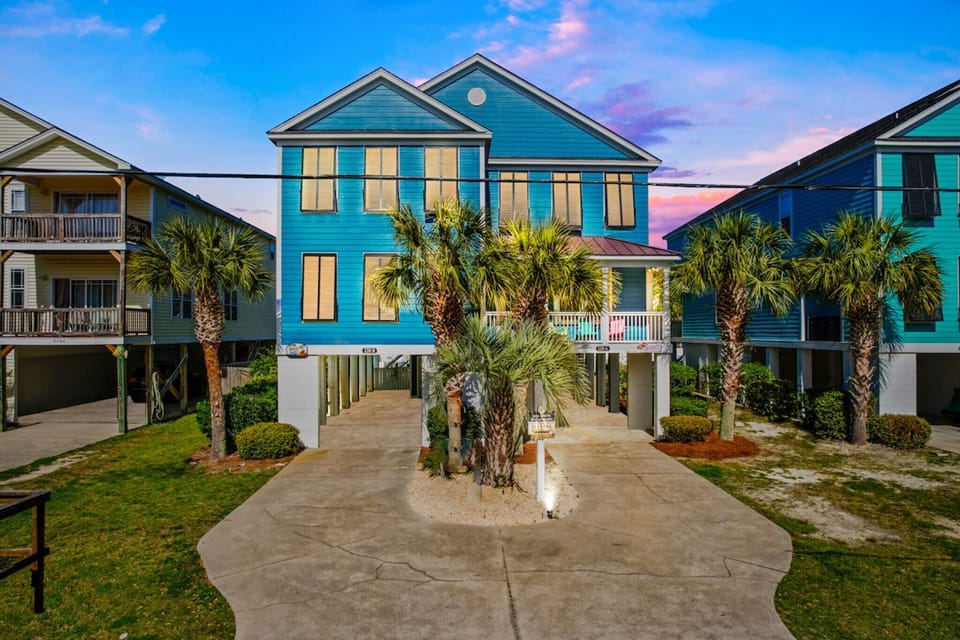 Vibrant blue beach house surrounded by swaying palms creates a tropical coastal retreat against stunning sunset skies.