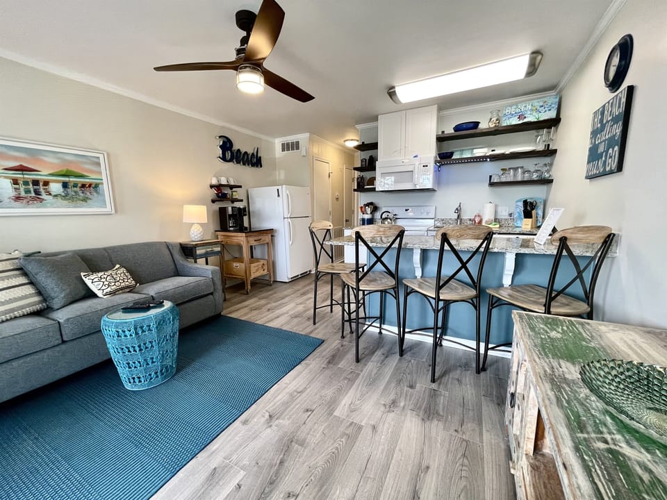 Compact living room and kitchen area with a gray sofa, blue rug, ceiling fan, bar stools, and coastal decor.