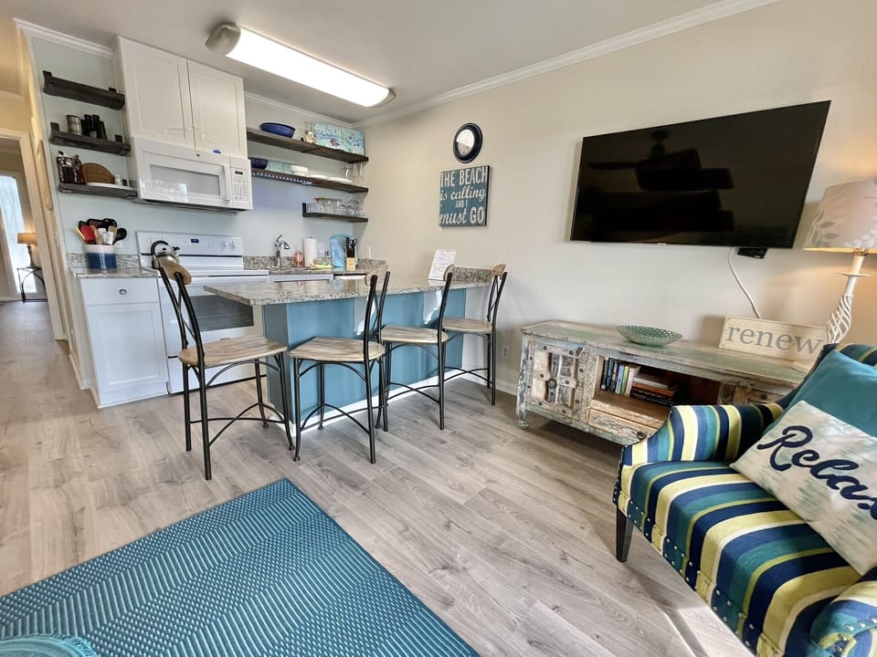Cozy kitchen and living area with bar stools, wall-mounted TV, striped chair, and beach-themed decor.