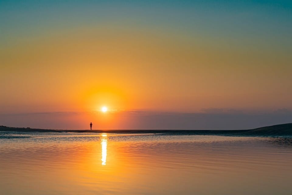 Person standing on a beach at sunset, with the sun low on the horizon, reflecting on the calm water.