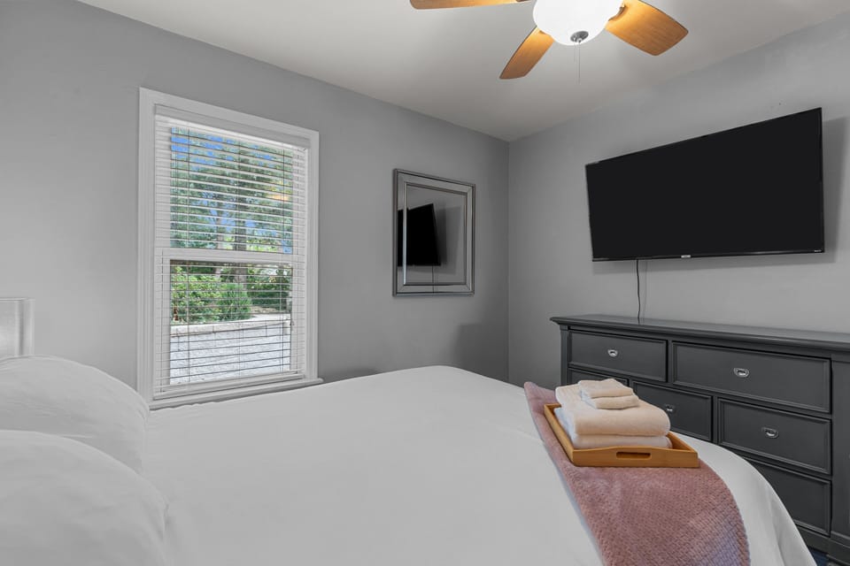King bedroom with a wall-mounted smart TV, bedside tables, ceiling fan, luggage bench, and fresh linens.