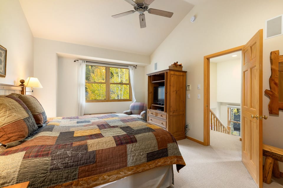 A bedroom with a quilted bed, wooden furniture, a ceiling fan, a window with a view of trees, and an open door showing a hallway and staircase.