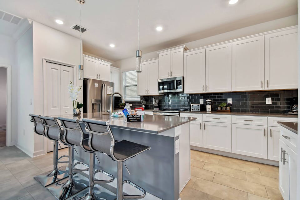 This bright kitchen boasts sleek white cabinetry and a spacious gray island ideal for dining or meal prep, while modern stainless steel appliances and stylish bar stools add a functional yet chic touch to the space.
