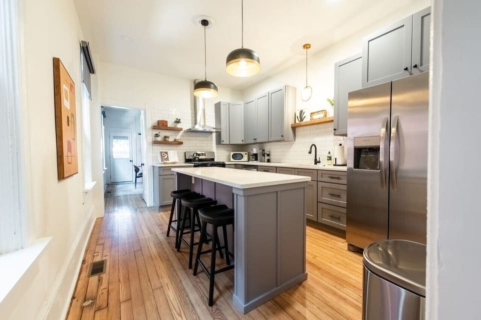 Modern kitchen with gray cabinets, a white island with black stools, stainless steel appliances, pendant lighting, and wooden floors
