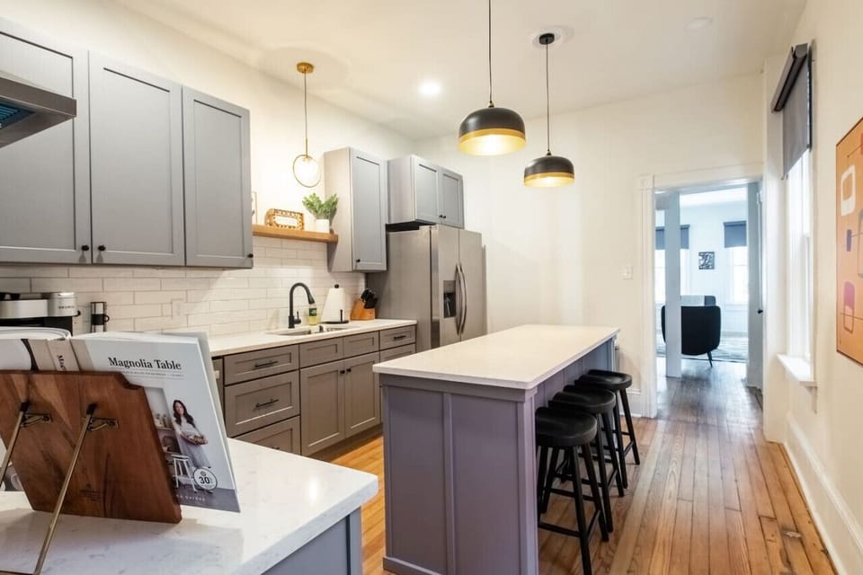 Modern kitchen with gray cabinets, a white island with black stools, stainless steel appliances, pendant lighting, and wooden floors