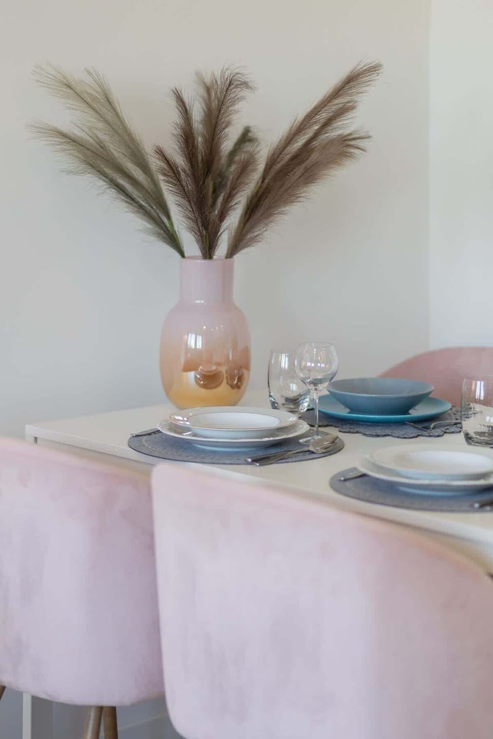 A pastel-themed dining table set with soft pink chairs, a vase with pampas grass, and elegant dinnerware.
