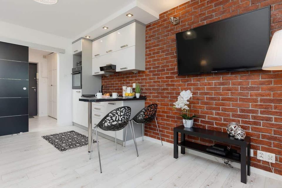 A living room with a flat-screen TV mounted on a red brick wall, a grey sofa, and a compact dining area.
