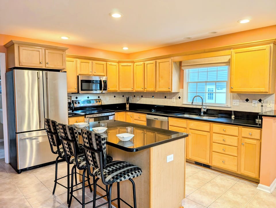 109 Soft Shell Cove - Modern Kitchen with Stainless Appliances - A perspective of the kitchen showcasing wooden cabinetry, black countertops, and stainless steel appliances, including a large refrigerator and microwave.