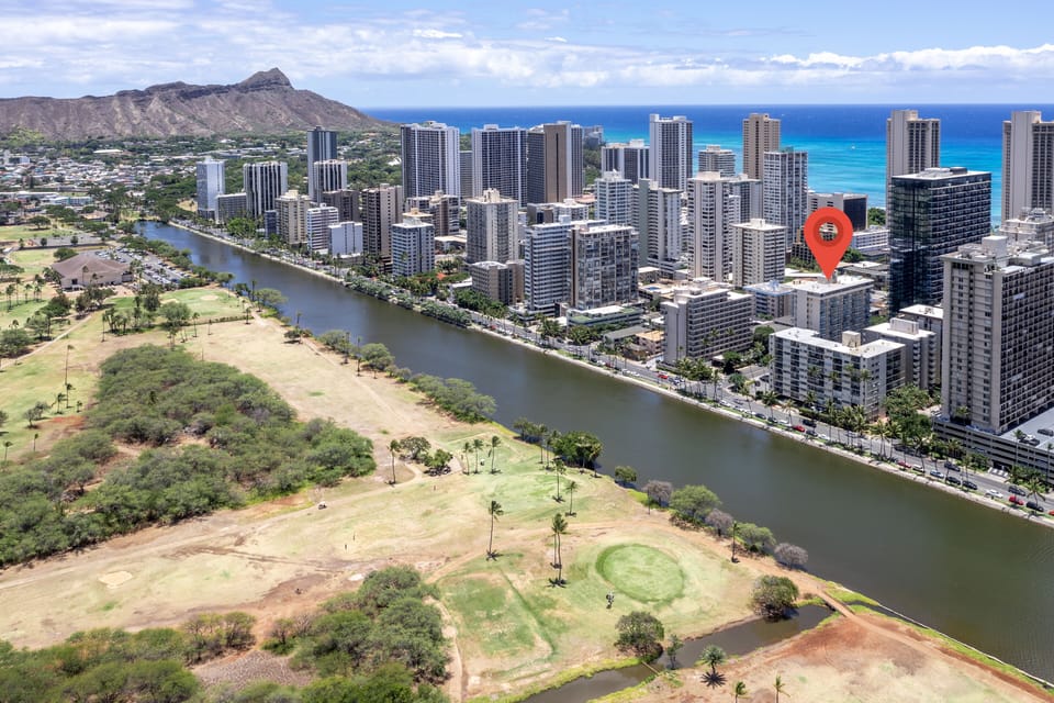Aerial view of a coastal urban area featuring high-rise buildings along a waterway, with Diamond Head crater and the Pacific Ocean in the background. Golf course and green spaces visible in the foreground.