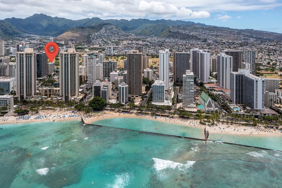 This aerial view captures the property's prime location along a stunning sandy beach, turquoise waters, and dramatic mountain backdrop in a vibrant urban setting.