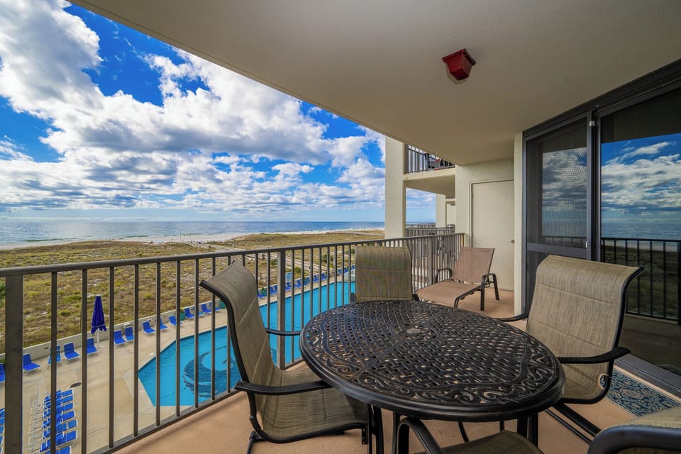 Balcony seating offers a view of the pool area and the beach, framed by a bright sky with scattered clouds