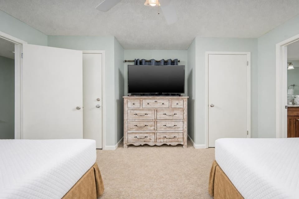 Guest bedroom features two double beds, a wooden dresser with a TV, and light green walls creating a calming atmosphere