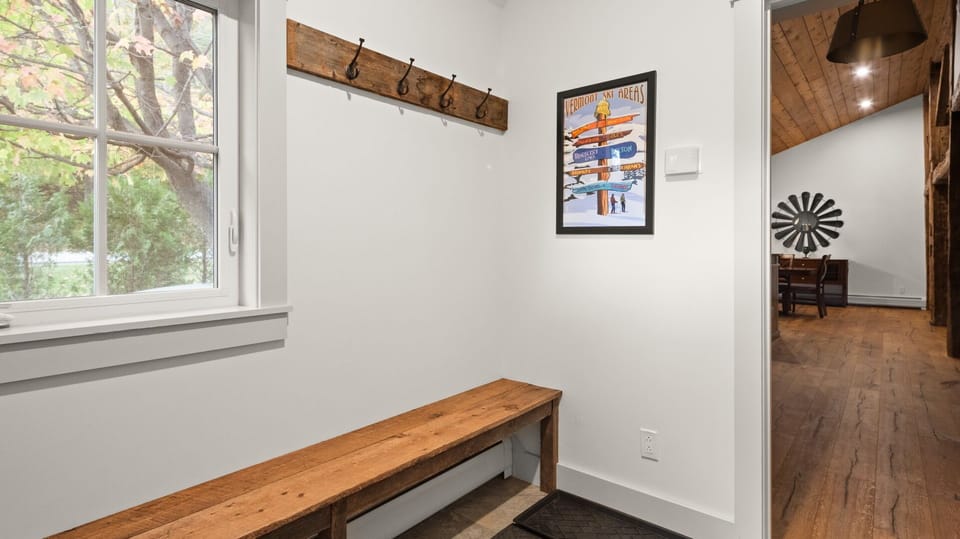 A small entryway with a wooden bench and coat hooks on the wall. A framed artwork of skis hangs next to a window. A hallway on the right leads to a kitchen area.