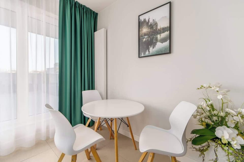 A minimalist dining area with a round table, white chairs, and a vase of flowers, located near a window with green curtains.
