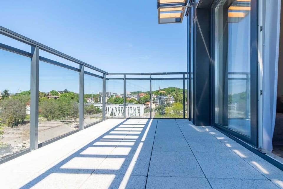 A spacious balcony with glass railings, offering a stunning view of the coastline and lush greenery.
