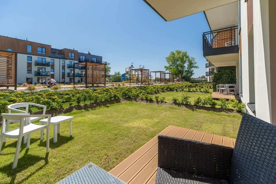 A sunny ground-floor terrace featuring a grassy area, outdoor seating, and neighboring apartment buildings in view.

