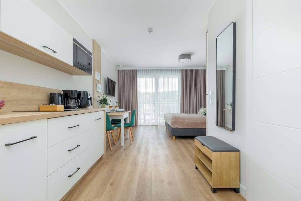 A modern kitchenette with white cabinets, a countertop, and built-in appliances. A wooden floor and a small seating area with a bench and table are visible further into the apartment.