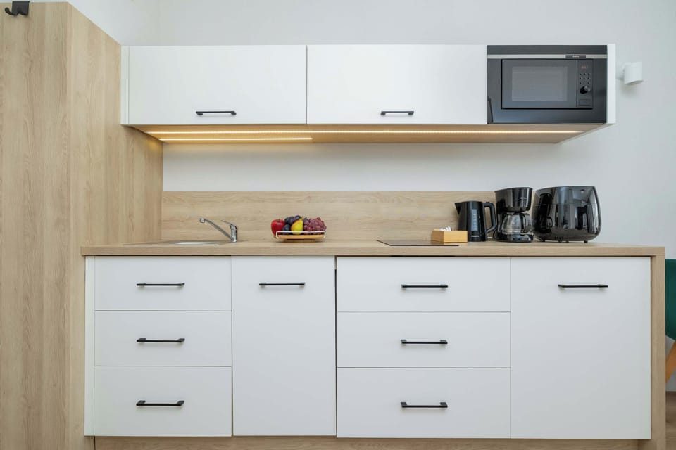 A modern kitchenette featuring white cabinets, a light wood countertop, and built-in appliances, including an oven and microwave. A coffee machine and other kitchen essentials are arranged on the counter.
