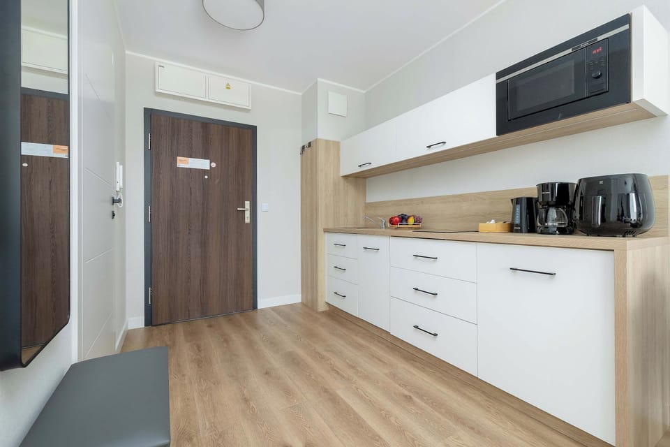 A bright apartment entrance with a dark wooden front door, white walls, and light wood flooring. A modern kitchenette with white cabinets and built-in appliances is visible to the right.