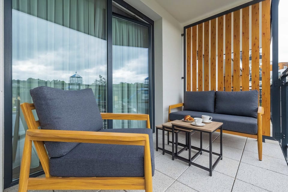A furnished balcony featuring comfortable blue and wooden seating, a small coffee table, and vertical wooden privacy panels. Large glass doors provide access to the apartment.