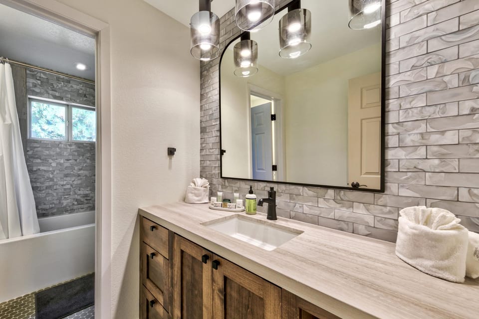 Bathroom vanity with wood cabinetry, light marble countertop, black faucet, and large arched mirror over a gray patterned tile backsplash.